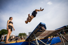 Mann springt rückwärts von einem Trampolin; links steht eine Frau, im Hintergrund stehen mehrere Personen.