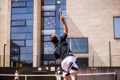 Tennisspieler schlägt den Ball über das Netz vor einem Gebäudekomplex.