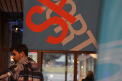Großes blaues Banner mit rotem und beigefarbenem Schriftzug; unten links ein Mann, rechts eine Person im Hintergrund.