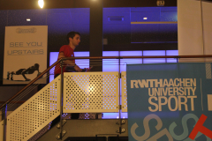 Person in rotem T-Shirt auf Treppe, daneben Banner „RWTHAACHEN UNIVERSITY SPORT“ und „SEE YOU UPSTAIRS“.
