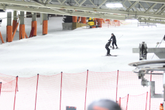 Skihalle mit zwei Skifahrern auf weißer Piste, rote Netze vorne und orange Stützen links.