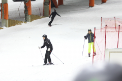 Drei Skifahrer auf einer schneebedeckten Piste, daneben ein rotes Sicherheitsnetz und orange Markierungen.