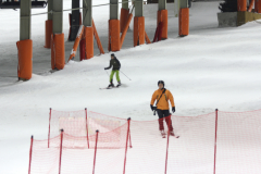 Zwei Skifahrer auf einer verschneiten Piste, unten rote Fangnetze, oben orange gepolsterte Stützen.