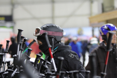 Person mit Helm, Skibrille und Gesichtsmaske zwischen vielen Skistöcken in einer Halle