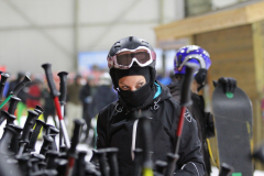 Person mit Skihelm und Skibrille zwischen mehreren Skistöcken und Snowboards in einer Halle.