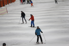 Mehrere Skifahrer auf einer schneebedeckten Piste; links ein rotes Netz, im Hintergrund weitere Personen.
