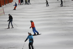 Mehrere Wintersportler fahren auf einer verschneiten Piste; links ein Skifahrer, mittig ein Snowboarder.