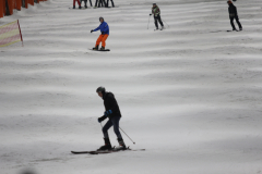 Mehrere Skifahrer und Snowboarder auf einer verschneiten Piste, links ein rotes Netz.