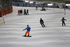 Mehrere Wintersportler auf einer beleuchteten Indoor-Skipiste, einige auf Skiern, einer auf dem Snowboard.