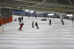 Mehrere Personen fahren auf Snowboards in einer Indoor-Skihalle auf einer verschneiten Piste.