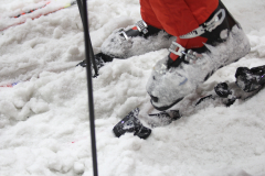 Nahaufnahme von Skischuhen mit Bindung im Schnee, daneben ein Skistock.