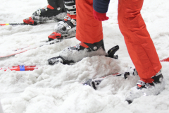 Skischuhe und rote Skihosen stehen im Schnee, rote Skier liegen teilweise sichtbar im Vordergrund.