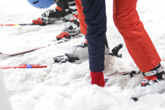Skifahrerbeine mit Skischuhen und Skiern im Schnee, rote und dunkelblaue Skihose, blaue Mütze.