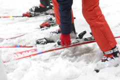 Personen mit Skischuhen und Skiern im Schnee, rote Hose und rote Handschuhe sichtbar.