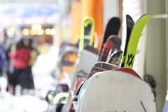 Mehrere Snowboards stehen aufrecht in einer Reihe; im Hintergrund unscharf stehende Personen und Lichter.