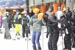 Gruppe von Ski- und Snowboardfahrern mit Helmen und Stöcken auf Schnee vor einem Gebäude.