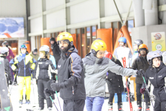 Mehrere Personen mit Skihelmen und Skistöcken stehen in einer Halle; einige tragen Skikleidung.
