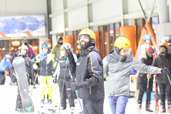 Personen mit Skikleidung, Helmen und Skistöcken in einer Halle, links ein Snowboard.