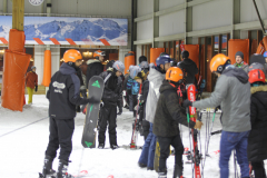 Gruppe von Skifahrern und Snowboardern in einer Skihalle mit Helmen, Skiern und Snowboards.