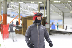 Person mit roter Mütze, grauer Jacke und schwarzen Handschuhen in einer Skihalle, andere Personen im Hintergrund.