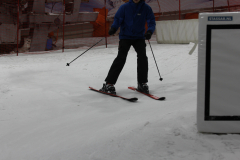 Skifahrer in blauer Jacke auf einer Indoor-Skipiste mit roten Netzen und weißem Schild rechts.
