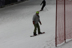 Snowboarder mit grünem Helm auf verschneiter Piste, rotes Netz rechts, Skifahrer im Hintergrund.
