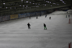 Indoor-Skihalle mit mehreren Skifahrern auf verschneiter Piste, Beleuchtung an der Decke, Schneehügel rechts.