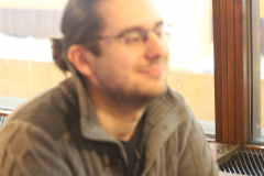 Unscharfes Porträt eines Mannes mit Brille und Zopf, seitlich vor einem Fenster sitzend.