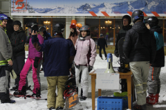 Gruppe von Schneesportlern mit Helmen auf Schnee vor einem Gebäude; Tisch mit Megafon und Schild.