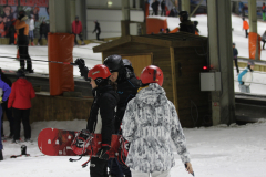 Drei Personen mit Helmen auf einer Skipiste; eine hält ein rotes Snowboard, im Hintergrund weitere Skifahrer.