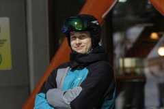 Person in Skikleidung mit Helm und Skibrille, Arme verschränkt, vor unscharfem Hintergrund.