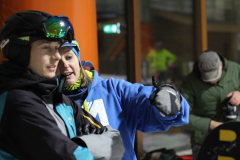 Person in blauem Hoodie und Helm mit ausgestrecktem Arm; links und rechts weitere Personen, eine hält eine Kamera.