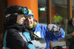 Zwei Personen in Winterkleidung mit Helmen, eine mit erhobenem, behandschuhtem Arm; Gesichter verpixelt.