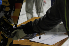 Handschuh greift ein Snowboard an einem Tisch; daneben liegen Karten und ein Blatt Papier mit Text.