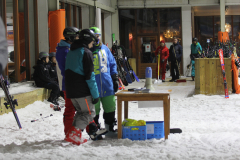 Skifahrer mit Helmen in einer verschneiten Halle, daneben Skier, ein Tisch und eine Kehre.
