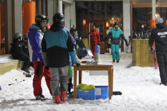 Mehrere Personen in Skikleidung in einem verschneiten Bereich mit Tisch, Ski und Skistöcken vor einem Gebäude.