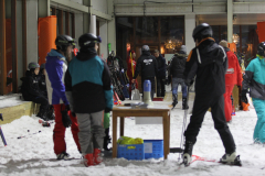 Mehrere Personen mit Ski- und Snowboardausrüstung stehen und sitzen in einer überdachten, verschneiten Halle.