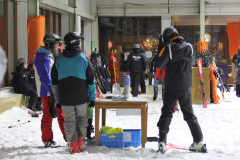 Menschen in Skikleidung in einer Skihalle, daneben Skier an den Wänden und ein Tisch im Schnee.