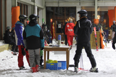 Mehrere Skifahrer mit Helmen stehen in einem verschneiten Innenbereich; Ski lehnen an Wänden und ein Tisch steht in der Mitte.