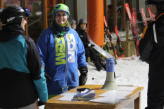 Mehrere Personen in Skikleidung stehen an einem Tisch mit Lautsprecher und Papieren, Ski im Hintergrund.