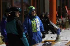 Mehrere Personen in Skihelm und Winterkleidung stehen im Schnee neben einem Tisch mit Megafon und Karten.