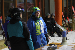 Personen mit Skihelmen und Winterkleidung stehen und sitzen vor einem Gebäude; Skier lehnen an der Wand.