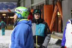 Personen mit Skikleidung und Helmen vor einem Gebäude; Schnee am Boden, eine Person im Vordergrund links.