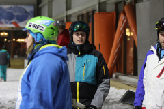 Mehrere Personen mit Skihelm und Wintersportjacken stehen in einer Halle mit Schnee auf dem Boden.