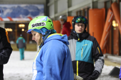 Personen mit Skihelmen und Winterjacken in einer Halle mit Schnee am Boden.