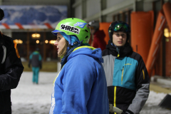Personen im Schnee tragen Helme und Winterjacken; im Hintergrund ein beleuchtetes Gebäude.