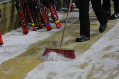 Person fegt Schnee mit einem Besen; links stehen mehrere rote Skier an der Wand.