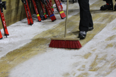 Person fegt Schnee mit Besen auf einer beschneiten Holzfläche; links stehen mehrere rote Skier.