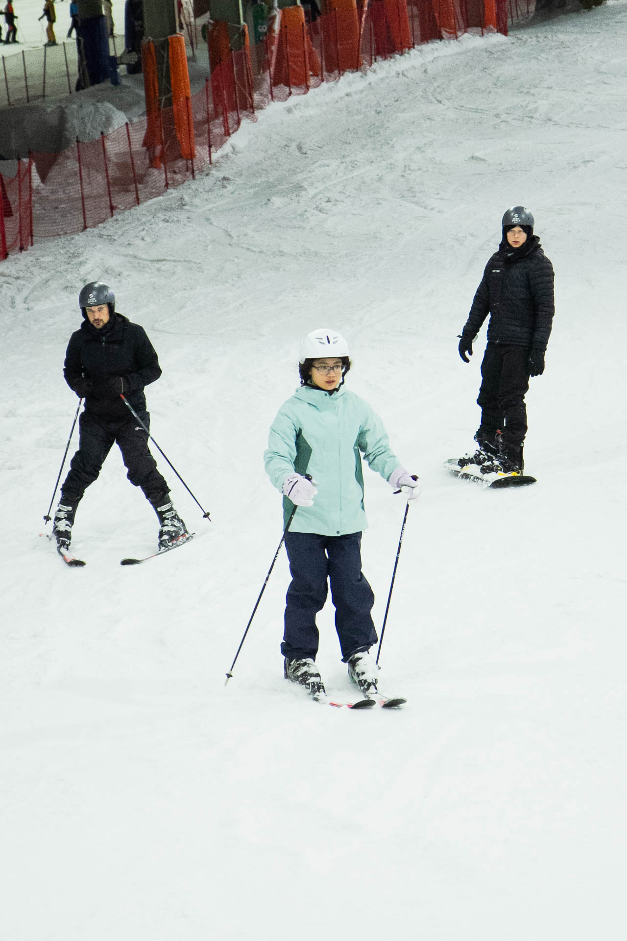 Drei Personen auf Skiern in winterlicher Umgebung mit einem ländlichen Gebäude und Zaun im Hintergrund.