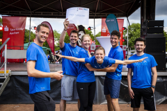 Sechs Menschen in blauen T-Shirts stehen auf einer Bühne; eine Person hält ein Schild hoch.